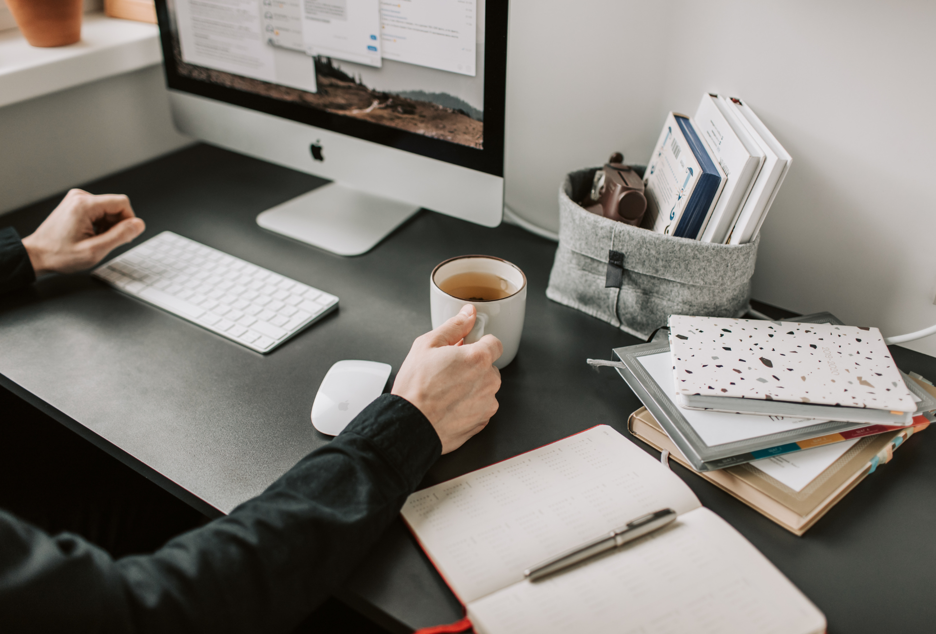 black desk and mac with a person holding a cup of tea and a stack of notebooks